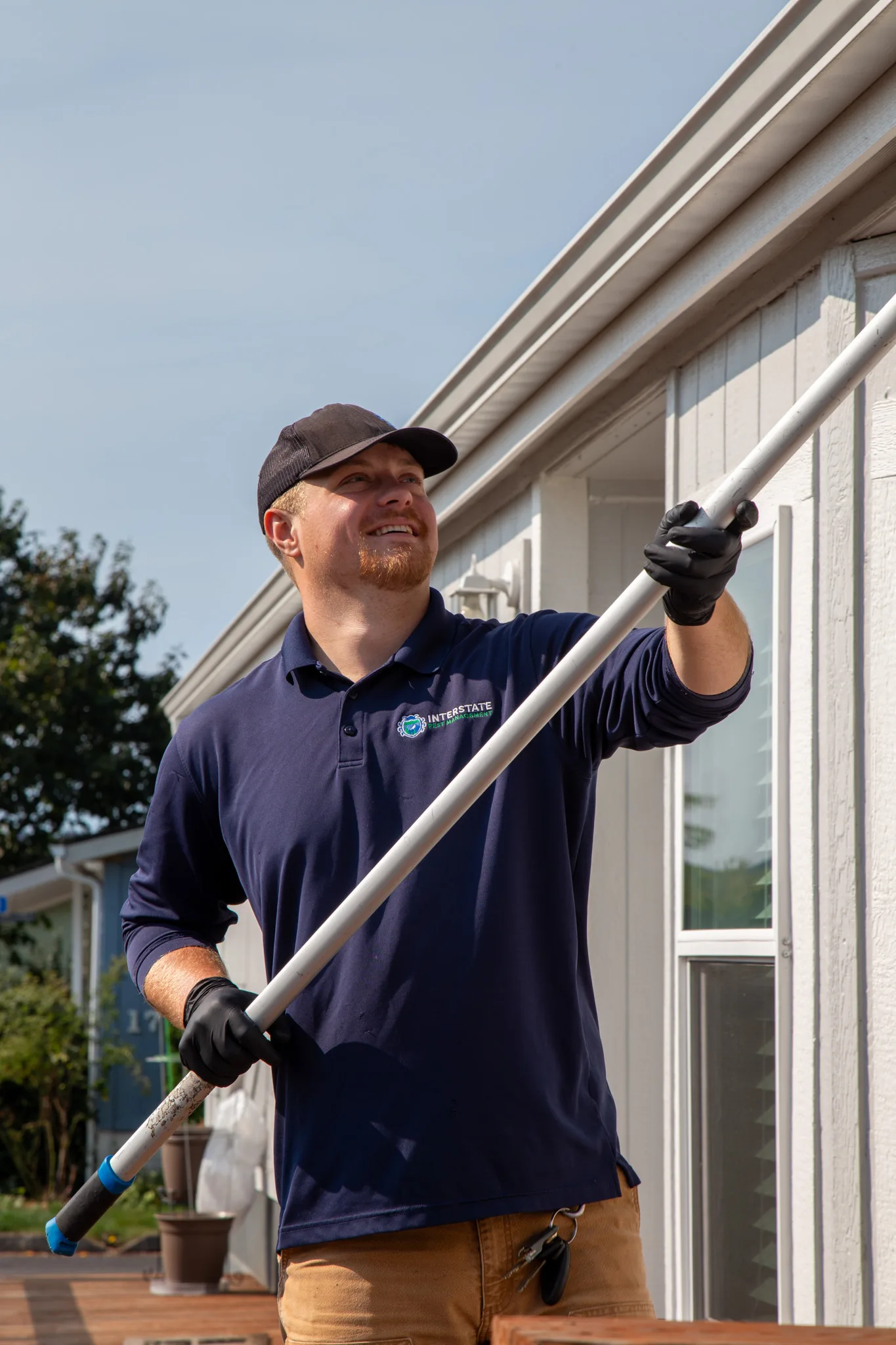 Professional pest control technician sweeping exterior house eaves to remove spider webs and insect nests